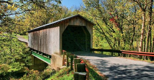 A wooden covered bridge spans a road, surrounded by vibrant green trees and a clear blue sky.