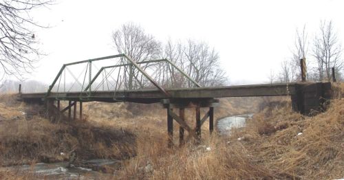 An old, green-trussed bridge spans a small creek, surrounded by dry grass and bare trees in a foggy landscape.