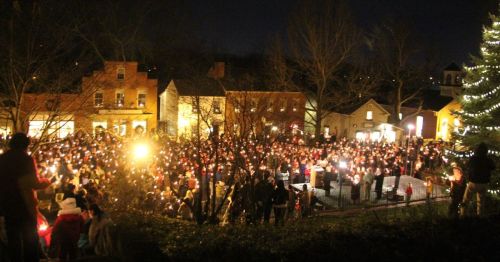 A large crowd gathers at night, holding candles near a decorated tree in a festive outdoor setting.