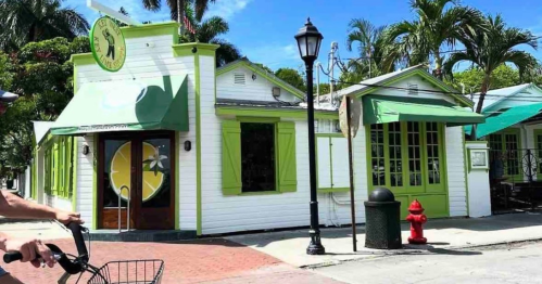 A colorful restaurant with green accents, palm trees, and outdoor seating on a sunny day. A cyclist passes by.