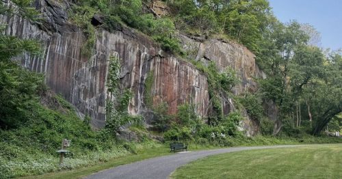 A scenic view of a rocky cliff with greenery, alongside a paved path and a bench in a peaceful outdoor setting.