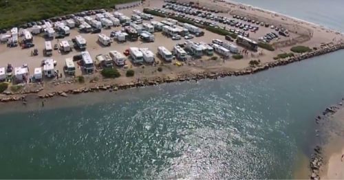 Aerial view of a beachside RV park with parked vehicles and a sandy shore along a calm, blue waterway.