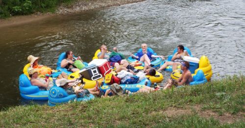 A group of people relaxes on colorful inflatable rafts in a river, enjoying drinks and each other's company.
