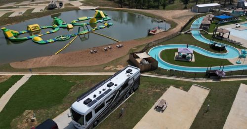 Aerial view of a recreational area with a pond, inflatable water park, and a winding water slide beside parked RVs.
