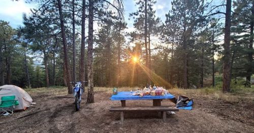 A campsite with a picnic table full of food, a tent, and a dirt bike, surrounded by tall trees at sunset.