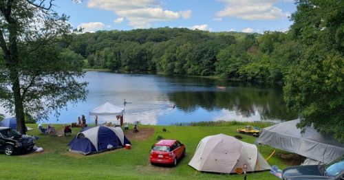 A serene lakeside scene with tents, a red car, and people enjoying the water under a blue sky with fluffy clouds.