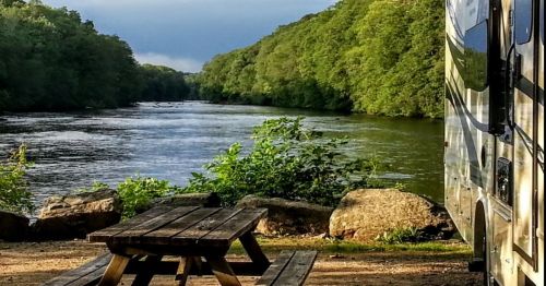 A serene river scene with lush greenery, a picnic table, and an RV parked nearby under a cloudy sky.