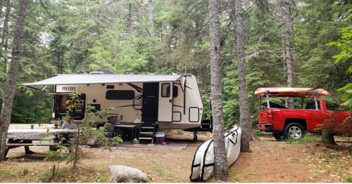 A camper and a red truck parked in a wooded area, surrounded by trees and outdoor gear.