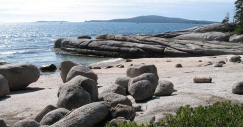 Rocky shoreline with smooth boulders, sandy beach, and calm water under a clear sky. Mountains in the distance.