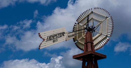 A vintage windmill with a wooden structure and blades, set against a blue sky with fluffy white clouds.