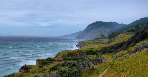 A scenic coastal view with rolling hills, cliffs, and waves crashing against the shore under a cloudy sky.