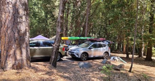 Two cars parked in a wooded area, with kayaks on top and camping gear scattered nearby.