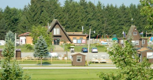 A scenic view of a campground with A-frame cabins, a main building, and playground equipment surrounded by trees.