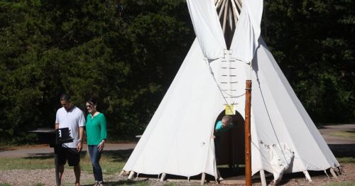 A couple stands near a teepee, while a child peeks out from inside, surrounded by trees and a pathway.