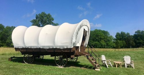 A covered wagon on wheels sits in a grassy field, with two white chairs and a small table nearby under a blue sky.
