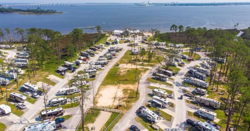 Aerial view of a large RV park by a lake, with numerous parked RVs and a bridge in the background.