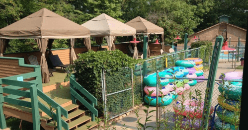 A pool area with shaded cabanas, colorful inner tubes, and a fenced-off swimming pool in a lush green setting.