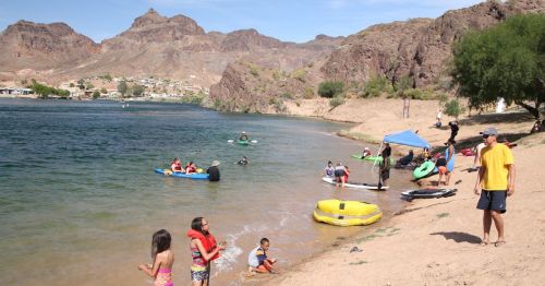 A sunny beach scene with people swimming, kayaking, and relaxing by the water, surrounded by rocky hills.