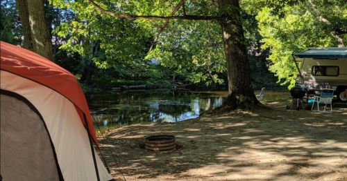 A campsite by a calm river, featuring a red tent, a camper, and green trees in a sunny setting.