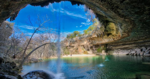 A serene cave with a waterfall cascading into a clear pool, surrounded by trees and rocky cliffs under a blue sky.