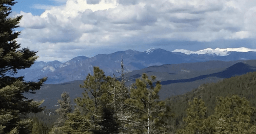 A scenic view of mountains with snow-capped peaks, surrounded by lush green trees and a cloudy sky.
