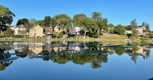 A serene lake reflects charming houses and trees under a clear blue sky.