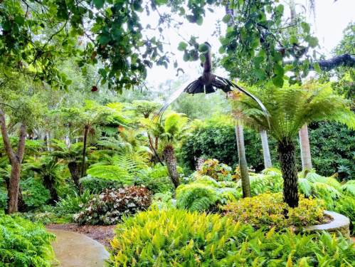Lush garden with ferns and tropical plants, featuring a pathway and a bird flying overhead amidst greenery.