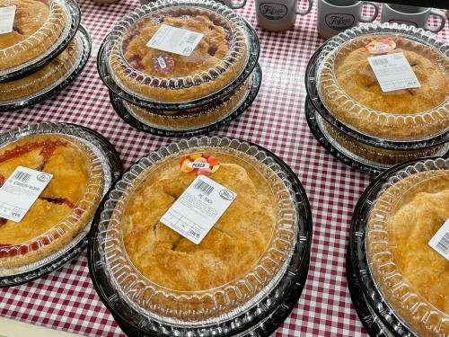 A display of various pies in clear plastic containers on a red and white checkered tablecloth.