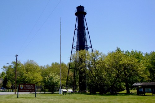 A tall black lighthouse stands beside a sign for Supawna Meadows National Wildlife Refuge, surrounded by green trees.