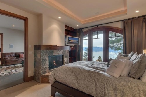Cozy bedroom with a stone fireplace, large windows, and a view of a lake and mountains. Soft bedding and warm lighting.