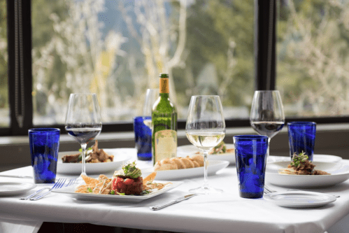 A beautifully set dining table with wine glasses, blue water cups, and gourmet dishes, with a scenic view outside.
