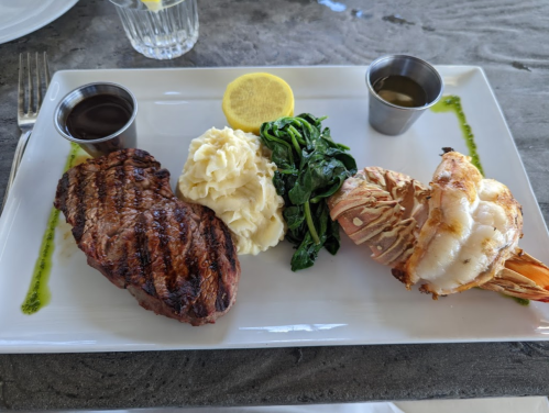A plate featuring grilled steak, shrimp, mashed potatoes, sautéed spinach, a lemon slice, and two dipping sauces.