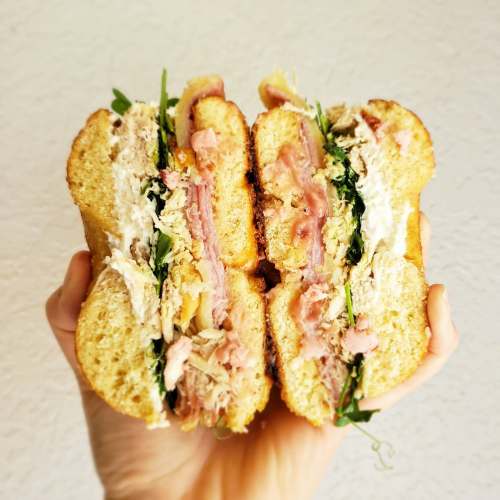 A hand holding a large sandwich filled with layers of meat, greens, and creamy spread, cut in half to show the filling.