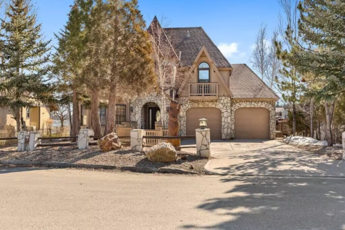 A large, stone-accented house with a sloped roof, surrounded by trees and a paved driveway.
