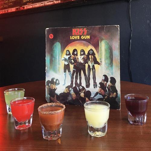 A KISS "Love Gun" album cover with six colorful shot glasses arranged in front on a wooden table.