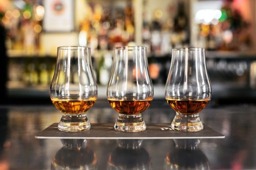 Three whiskey glasses filled with amber liquid, placed on a black mat against a blurred bar background.