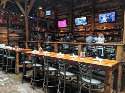 A rustic restaurant interior with wooden tables, bar stools, and multiple TVs showing sports. Two patrons are seated at the bar.