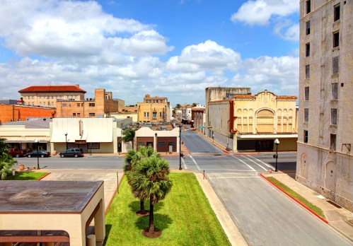 A sunny street view of a small town with historic buildings, palm trees, and a blue sky with fluffy clouds.
