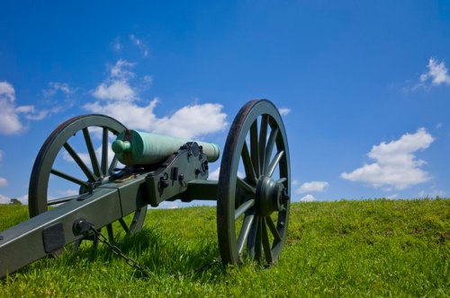 A historic cannon on a grassy field under a bright blue sky with fluffy white clouds.