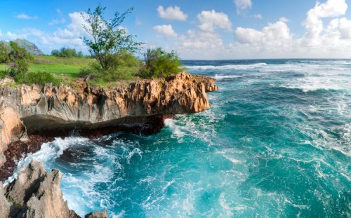 A rocky coastline with turquoise waves crashing against cliffs under a bright blue sky with scattered clouds.