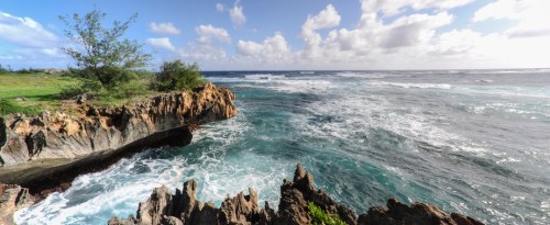 A panoramic view of rocky coastline with waves crashing against the shore under a bright blue sky with scattered clouds.
