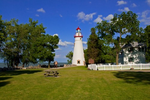 A white lighthouse with a red top stands near a grassy area, surrounded by trees and a blue sky with clouds.
