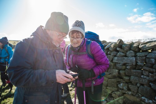 Two hikers, a man and a woman, smile as they look at a smartphone outdoors, with a stone wall and bright sunlight behind them.