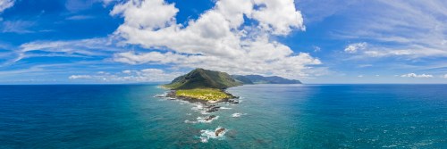 A panoramic view of a lush green coastline meeting a vibrant blue ocean under a partly cloudy sky.