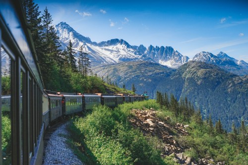 A scenic train travels along a mountain path, surrounded by lush greenery and snow-capped peaks under a clear blue sky.