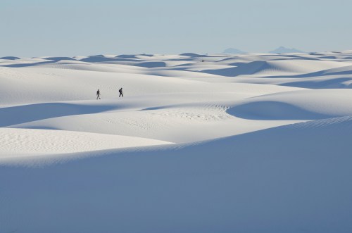 Two hikers traverse a vast, undulating landscape of white sand dunes under a clear blue sky.