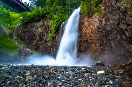 A cascading waterfall flows down rocky cliffs, surrounded by lush greenery and mist rising from the water.