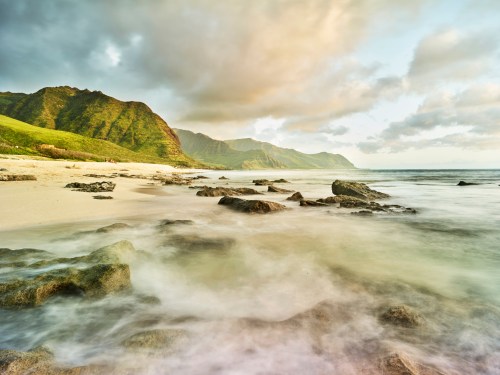 A serene beach scene with gentle waves, rocky shore, and lush green mountains under a cloudy sky.