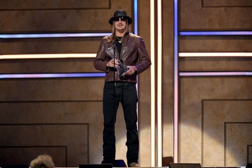 A man in a leather jacket and hat stands on stage holding an award, with colorful lights in the background.