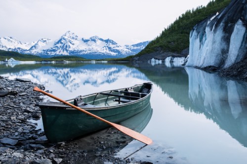 A green canoe with an orange paddle rests on a rocky shore, reflecting mountains and glaciers in calm water.